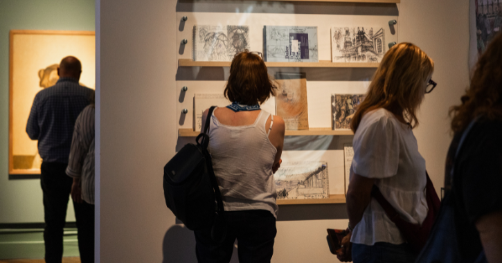 Photograph showing a group of people observing and discussing a framed artwork of a woman, displayed on a green wall in a gallery or museum setting. The artwork features warm tones and detailed shading, with accompanying text on the wall providing context about the piece and its significance.